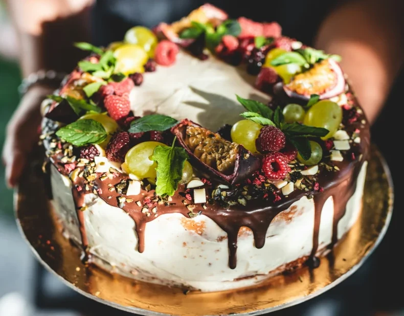 A person holding a white cake topped with chocolate and fresh fruit